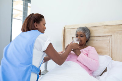 Female doctor giving glass of water to sick senior women on bed in bedroom